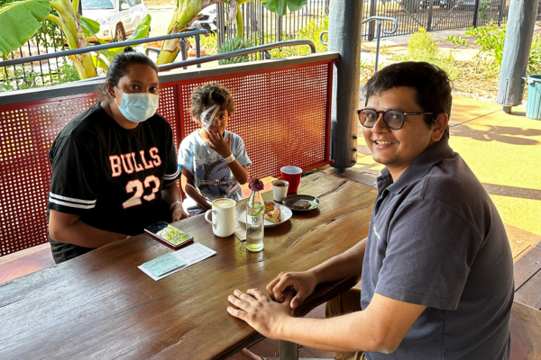 Joaquin sitting at an outdoor wooden table in Broome with his mum and Dr Viabhav Shah. Joaquin has a large patch over his right eye. Banana palms can be seen in the background.