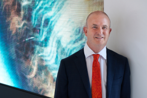 A head and shoulders photo of Professor Morgan standing in front of a large LED screen displaying an image of an eye. He is wearing a vibrant red tie, white collared shirt and navy suit jacket, and is smiling directly at the camera.